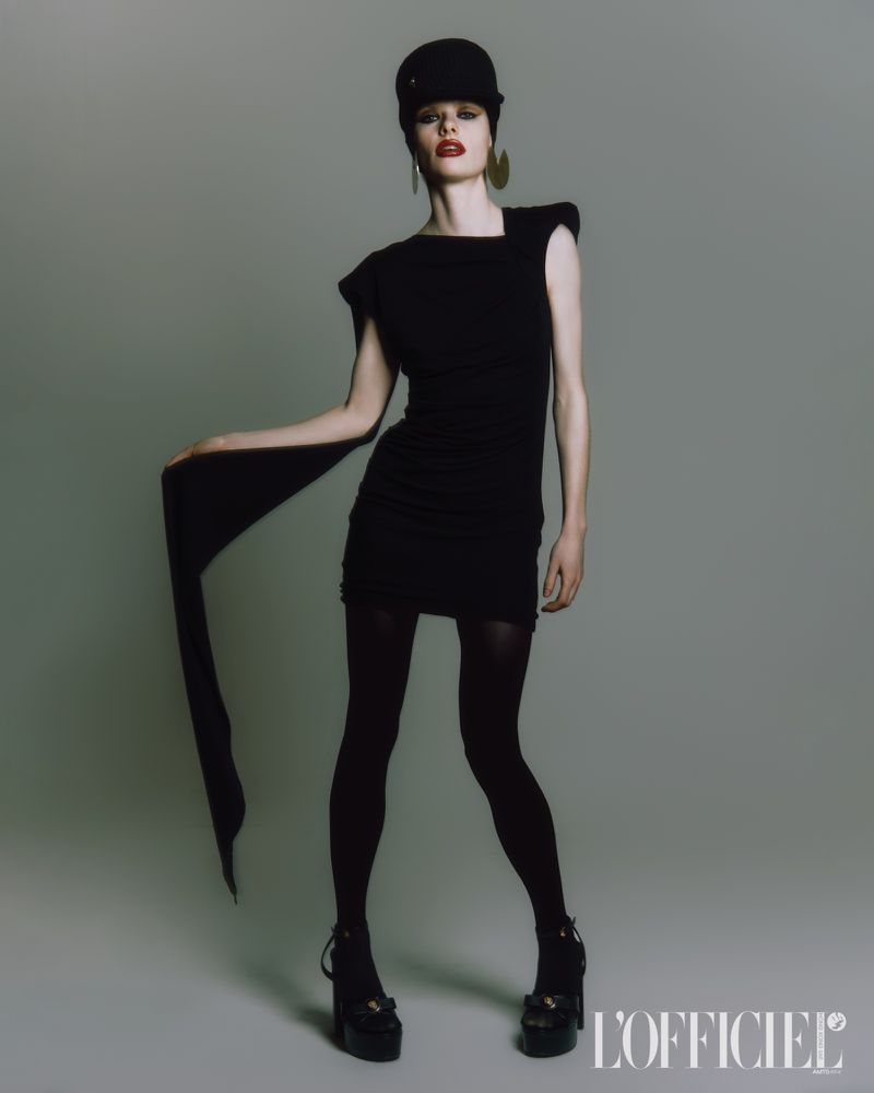 Full-length portrait of a woman posing in a black mini dress, cap, earrings, tights, and platform heels against a gray backdrop.