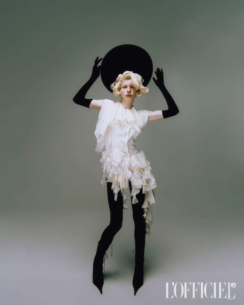 An adult female model poses in a short white dress with ruffles, black tights and boots, and long black gloves, holding a round black hat.