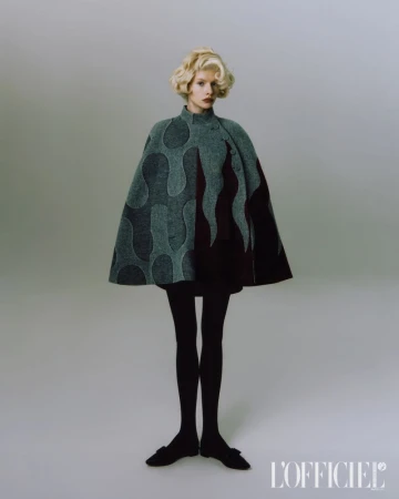 Model with blonde hair wearing a patterned grey cape and black tights against a grey seamless background