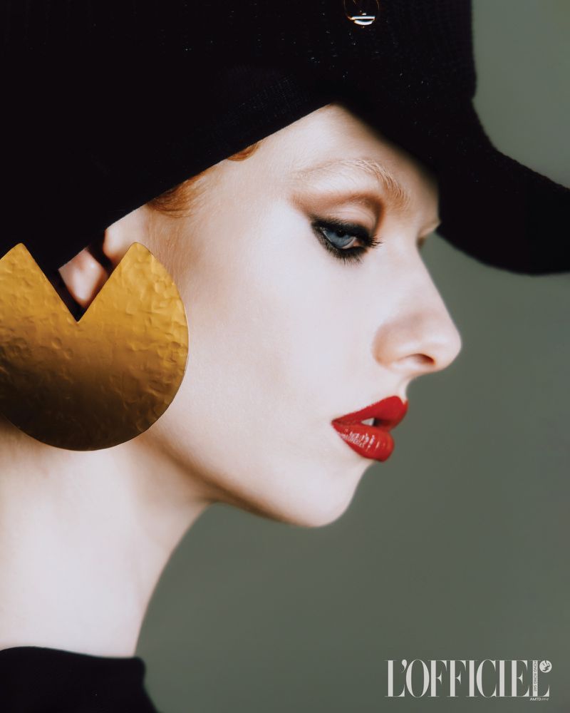 Portrait of a model wearing a black hat, large gold earring, and red lipstick in a studio with a grey background, showcasing beauty and fashion.