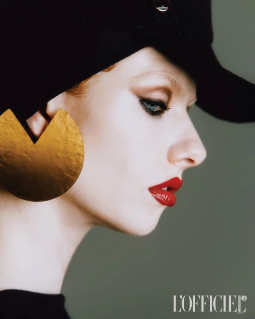 Portrait of a model wearing a black hat, large gold earring, and red lipstick in a studio with a grey background, showcasing beauty and fashion.