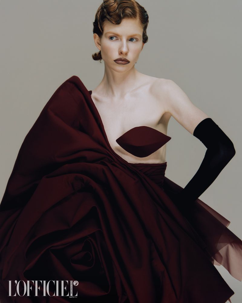 Fashion editorial shot of a woman wearing a dramatic maroon dress with a large bow, black gloves, and her hair styled in waves.