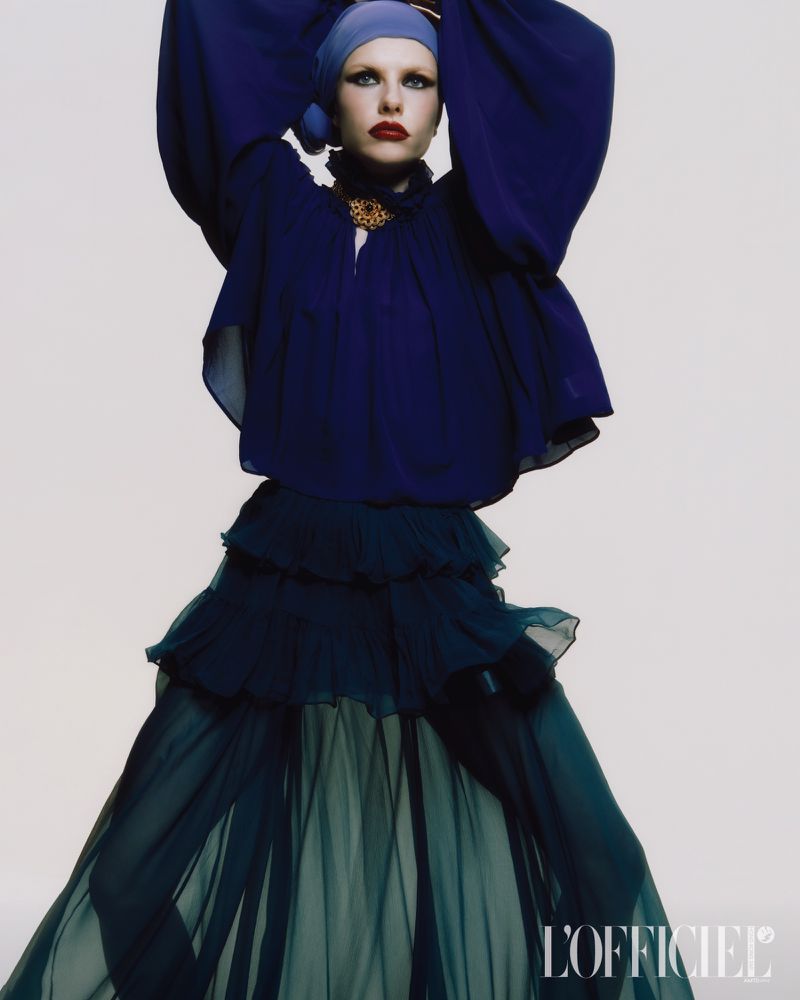 A fair-skinned woman posing in a blue headscarf, with a matching navy ruffled top, and a tiered, flowing skirt against a white backdrop.