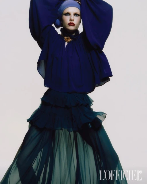 A fair-skinned woman posing in a blue headscarf, with a matching navy ruffled top, and a tiered, flowing skirt against a white backdrop.