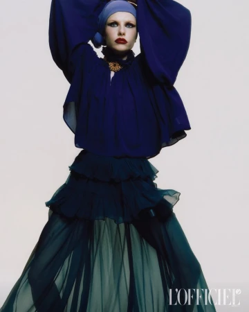 A fair-skinned woman posing in a blue headscarf, with a matching navy ruffled top, and a tiered, flowing skirt against a white backdrop.