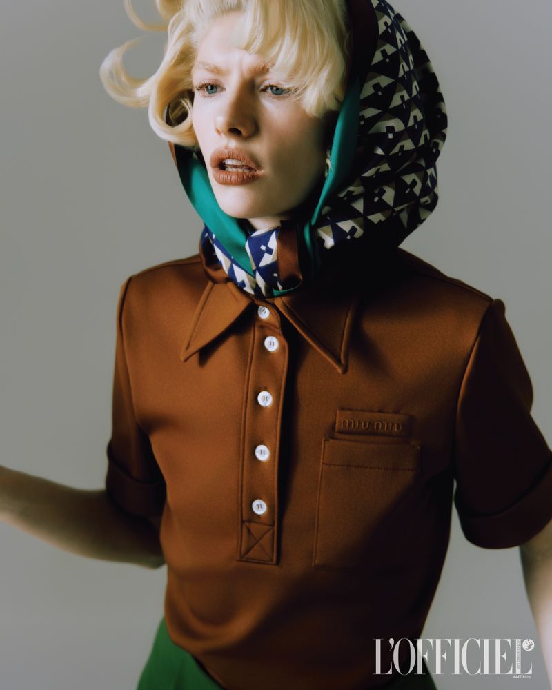 Close-up fashion shot of a blonde woman wearing a brown polo shirt, head scarf, and green pants against a grey backdrop in studio lighting.