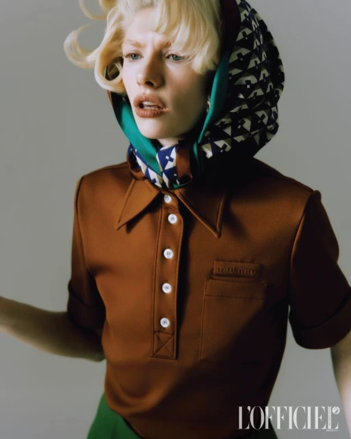 Close-up fashion shot of a blonde woman wearing a brown polo shirt, head scarf, and green pants against a grey backdrop in studio lighting.