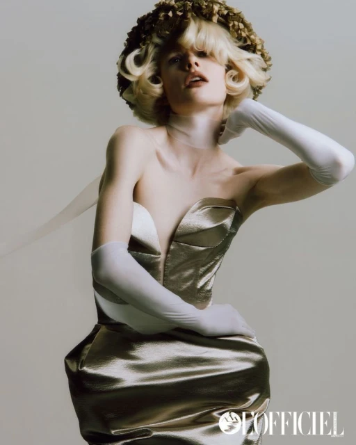 Model in gold strapless dress with flower crown and sheer gloves poses in a studio against a neutral background in portrait orientation.