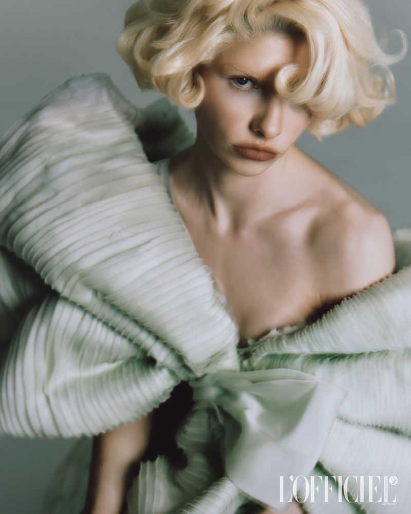 Fashion portrait of a fair-skinned model with blonde curly hair wearing a light green textured garment with large bow-like details.