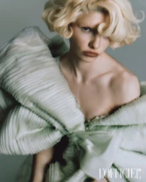 Close-up portrait of a blonde woman wearing a sculptural pale green ruffled dress against a grey background.
