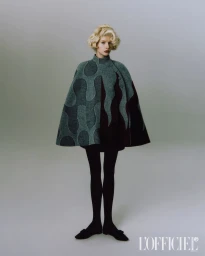 Model with blonde hair wearing a patterned grey cape and black tights against a grey seamless background