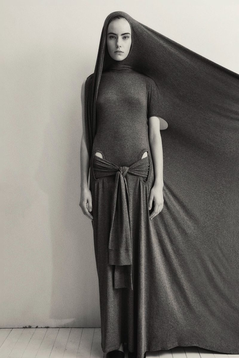 Monochrome fashion shot: A model in a grey draped gown with a hood poses against a neutral background in a studio setting.
