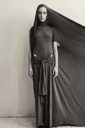 Monochrome fashion shot: A model in a grey draped gown with a hood poses against a neutral background in a studio setting.