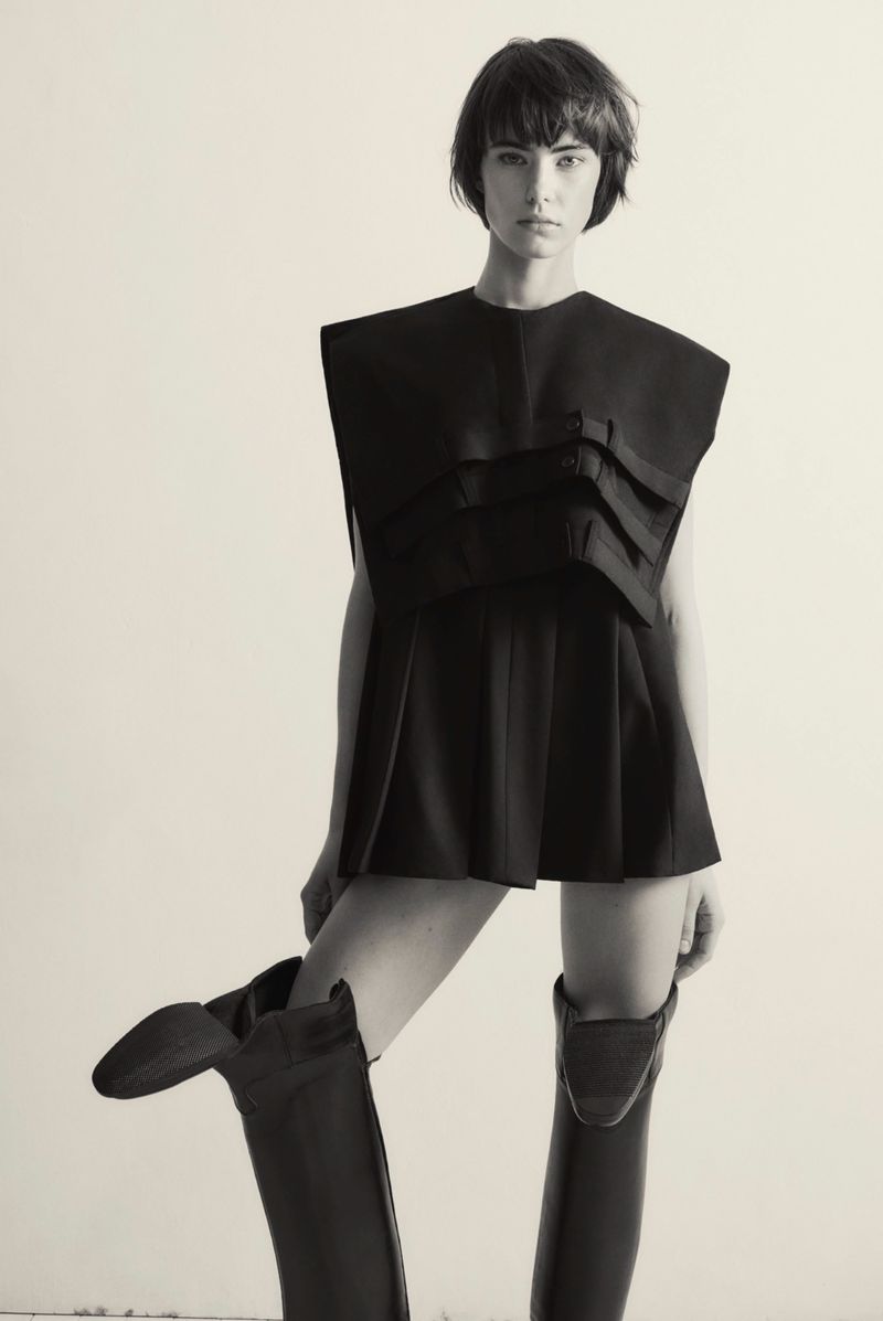 Monochromatic fashion portrait of a female model in a dark outfit with knee-high boots against a neutral background in a studio setting.