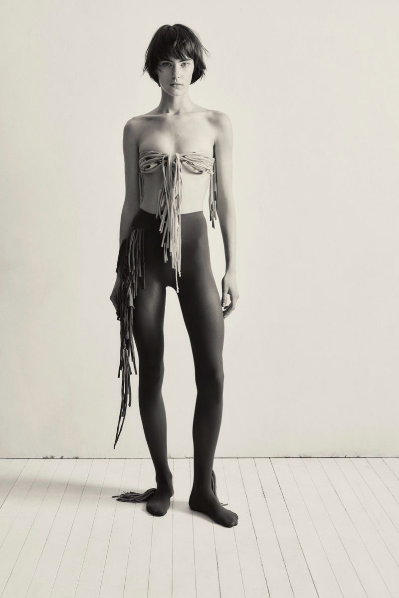 A woman stands in a photo studio wearing a fringed top and leggings, striking a pose against a neutral wall and wooden floor.