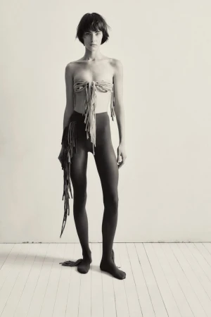 A woman stands in a photo studio wearing a fringed top and leggings, striking a pose against a neutral wall and wooden floor.