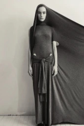 Model in a long grey draped hooded dress with a tied waist against a plain background