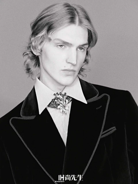 Portrait of a young adult male in a black velvet blazer with jeweled brooch, captured in black and white with soft, diffused lighting.