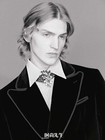 Portrait of a young adult male in a black velvet blazer with jeweled brooch, captured in black and white with soft, diffused lighting.
