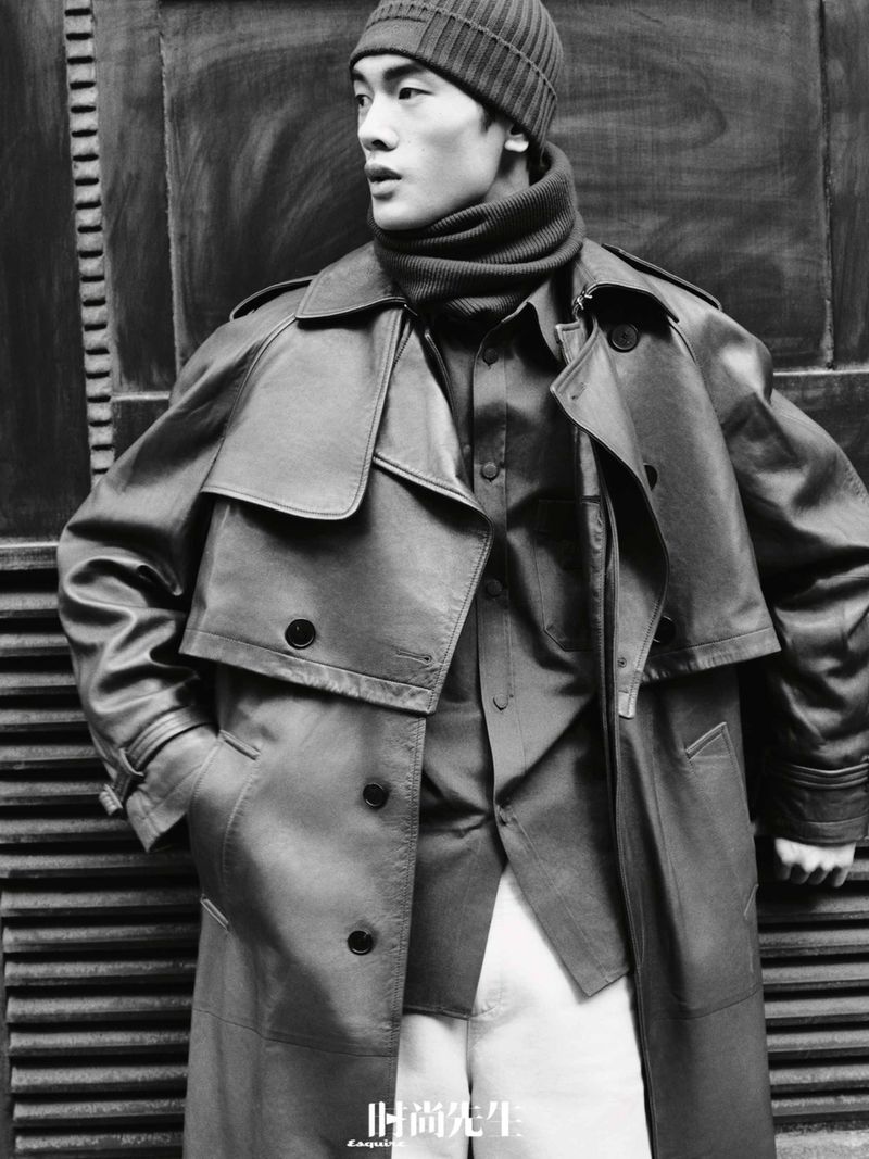 Monochrome fashion shot of an adult male in a trench coat, beanie, and scarf, standing against a grooved wall, fashion editorial.