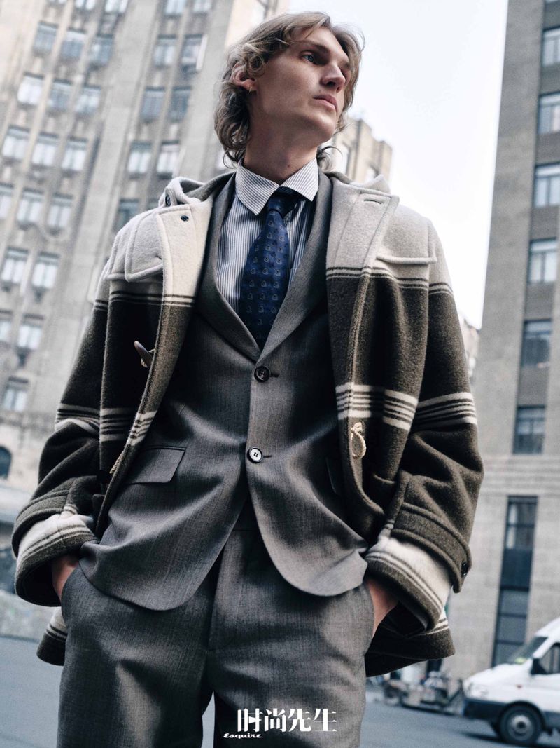 Male fashion model posing in a gray suit and striped hooded coat in a city environment with a tall building in the background.