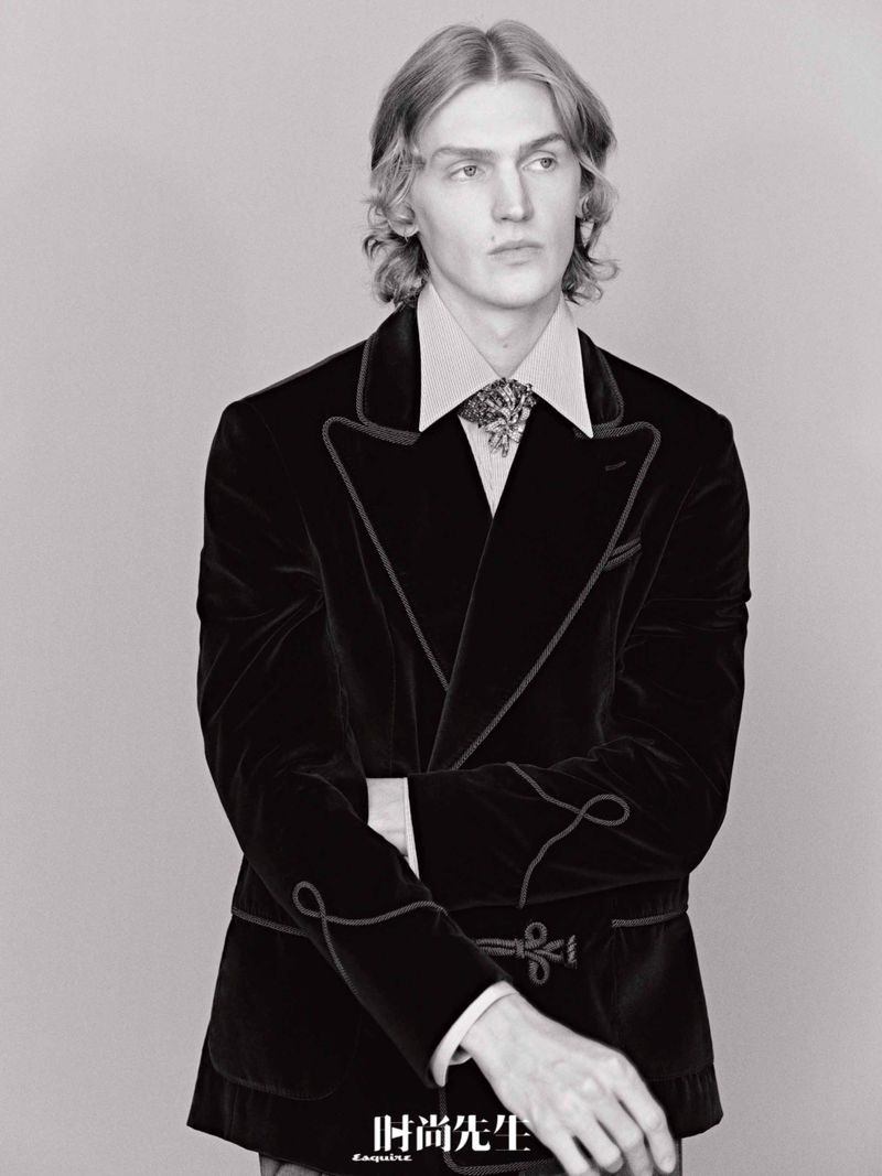 Fashion shot of a blonde male model wearing a black velvet blazer and collared shirt in a studio with gray backdrop, posing elegantly.