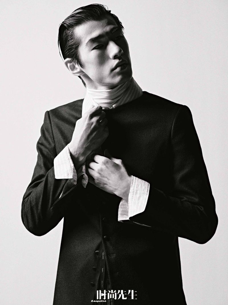 Black and white portrait of an Asian male fashion model in a dark suit adjusting his collar, set against a bright studio background.