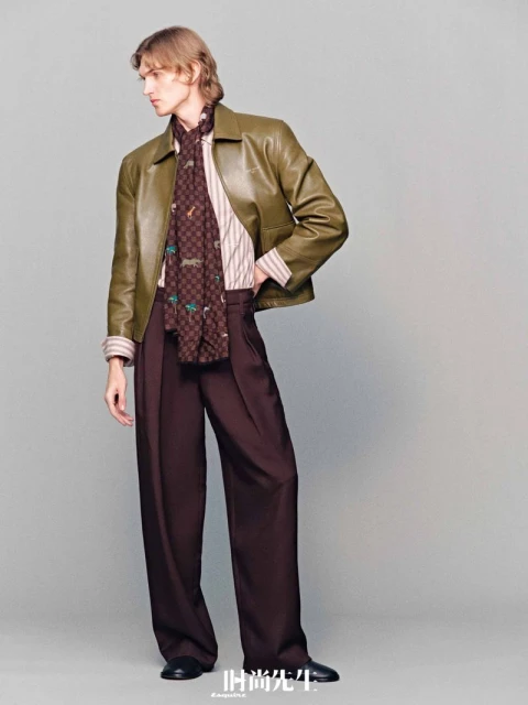 Full-length studio shot of a blond male model in olive leather jacket, striped shirt, brown scarf, and wide leg brown pants.