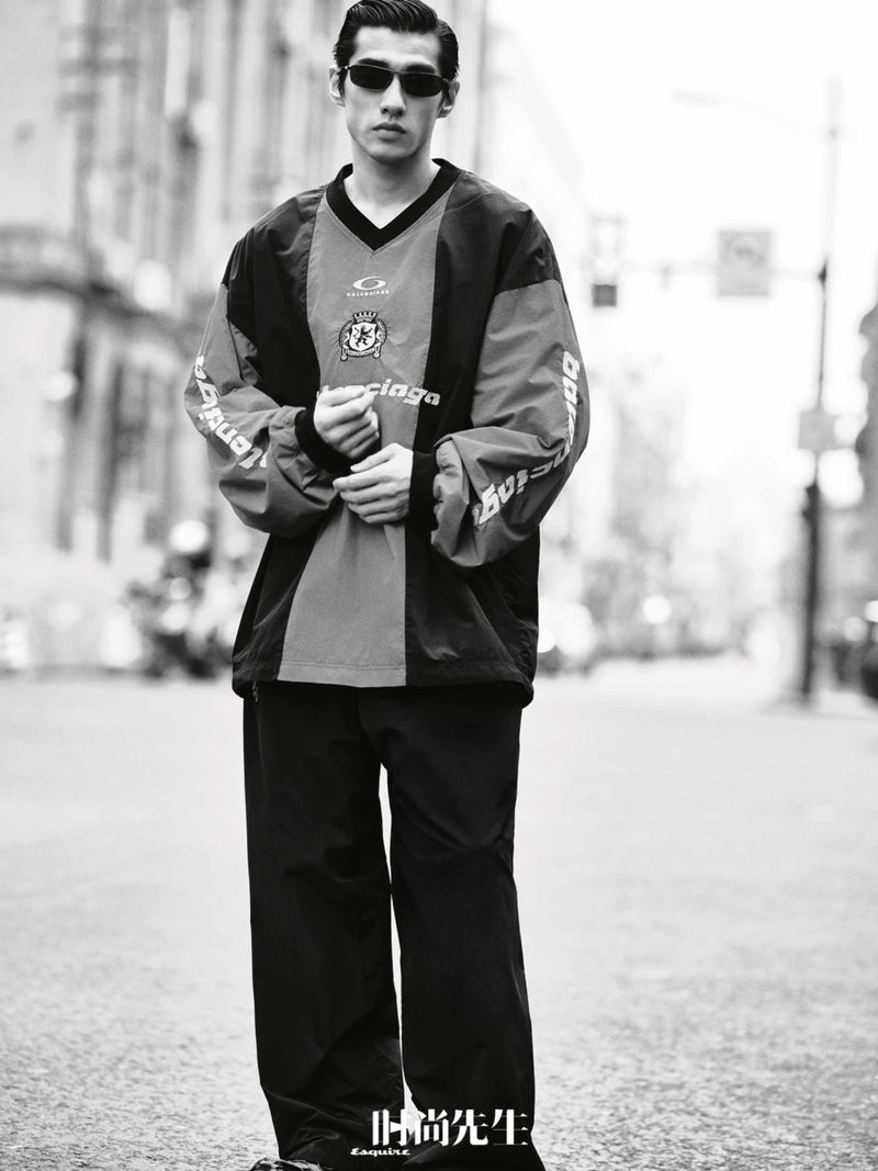 Monochrome portrait of Asian adult male in a sport-inspired fashion jacket and sunglasses, posing on a city street.