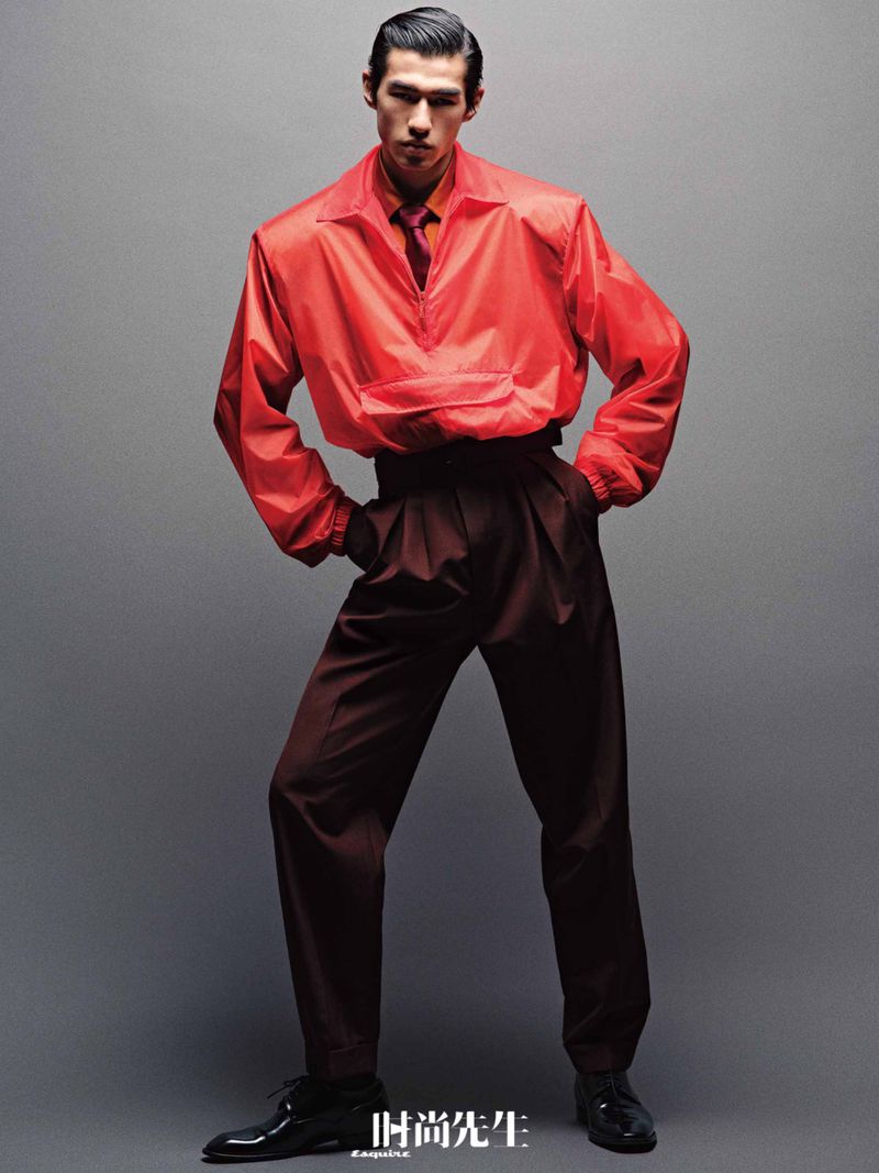 Fashionable Asian man poses in orange jacket and pleated trousers against a neutral backdrop, showcasing modern menswear style.