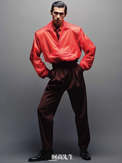 Fashionable Asian man poses in orange jacket and pleated trousers against a neutral backdrop, showcasing modern menswear style.