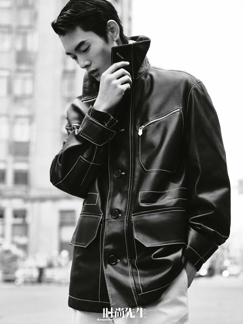 Monochrome medium shot of a young adult man in a black jacket posing outdoors with city buildings in the background, fashion editorial.