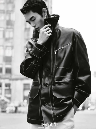 Monochrome medium shot of a young adult man in a black jacket posing outdoors with city buildings in the background, fashion editorial.