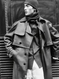 Monochrome portrait of an East Asian male model wearing a trench coat, turtleneck, and beanie against a dark architectural background.
