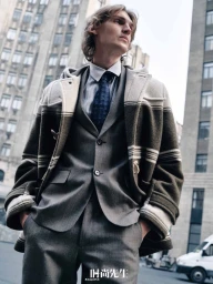 Model in a grey suit and patterned duffle coat standing outdoors in front of urban architecture