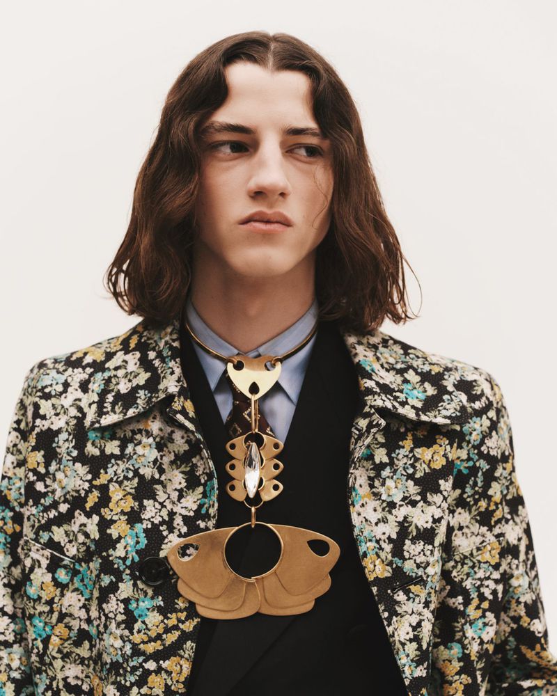 Male model in floral jacket with statement necklace poses in a studio with neutral backdrop, captured in a medium portrait shot.