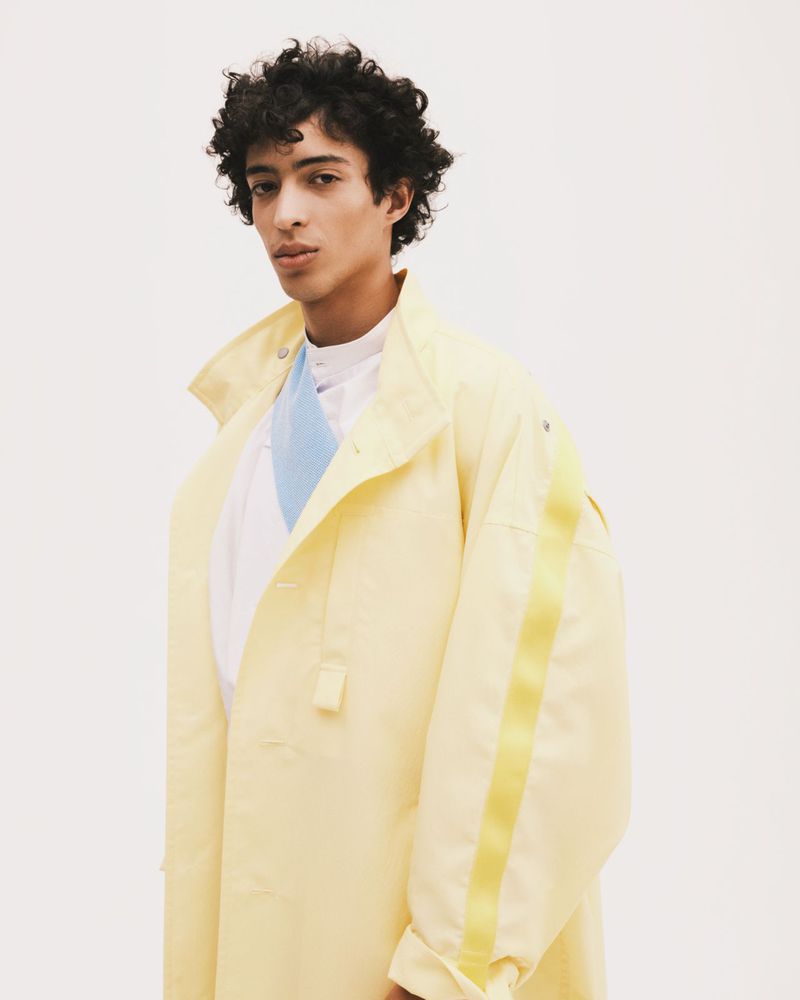 A young adult male poses in a studio wearing a pale yellow overcoat with a pale yellow stripe against a bright white backdrop.