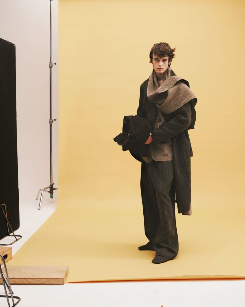 A fair-skinned young adult man in dark casual suit with scarf poses in a fashion shot in front of a pale-yellow backdrop in a photo studio.
