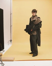 Model in dark wool coat and scarf holding a black bag standing against a yellow studio backdrop