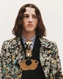 Close-up portrait of a male model with long wavy brown hair wearing a floral patterned jacket and a prominent gold metallic pendant necklace.