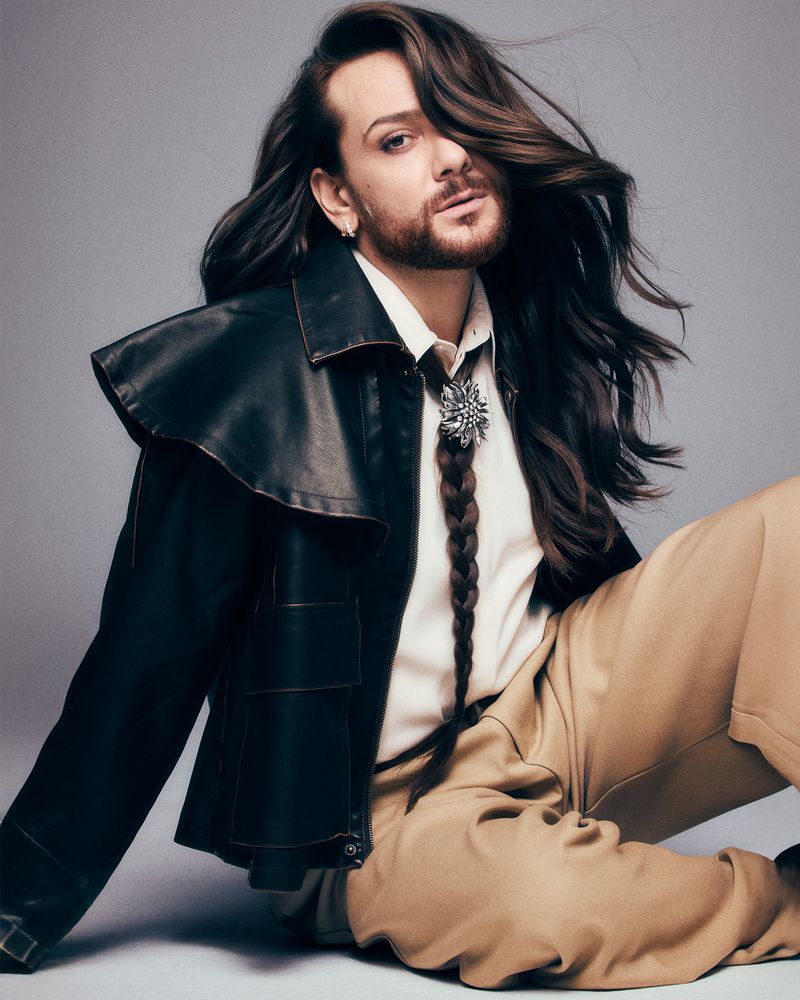 Portrait of an adult male with long brown hair and a beard, wearing a black leather jacket, off-white shirt and tan pants, posing indoors.