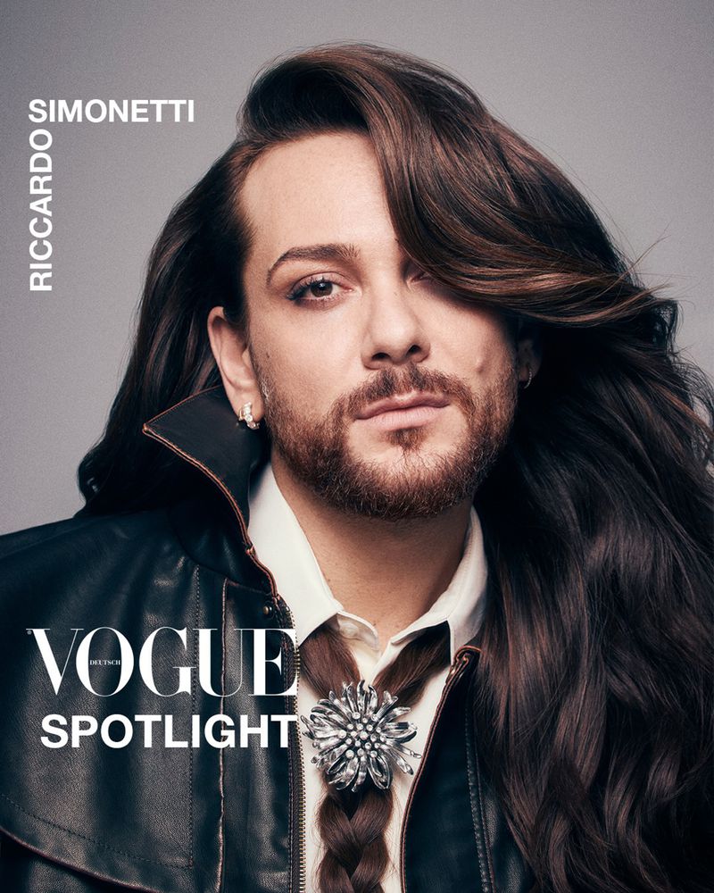 Portrait of Riccardo Simonetti with long brown hair, wearing a black leather jacket and decorative brooch for Vogue Spotlight.