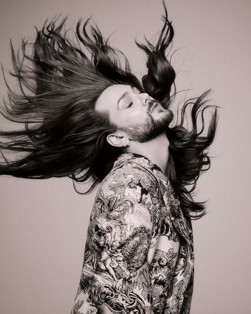 Portrait of a stylish adult male with long hair and a beard, wearing a patterned jacket, in a monochrome studio setting.