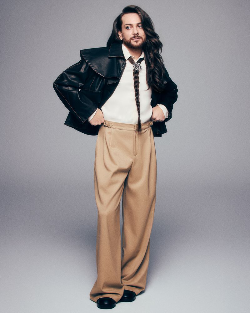 Man with long brown hair and a braided lock poses in a studio wearing a black leather jacket, cream sweater, and khaki wide-leg pants.