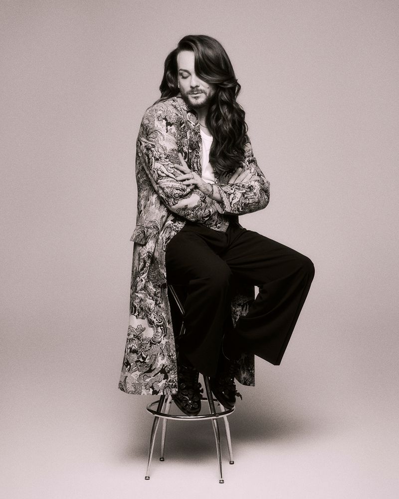 Portrait of a person with long hair wearing patterned coat and dark trousers, seated on a stool against a light grey backdrop.
