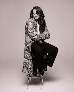 Riccardo Simonetti with long dark hair seated on a stool wearing a patterned coat and black trousers against a neutral background