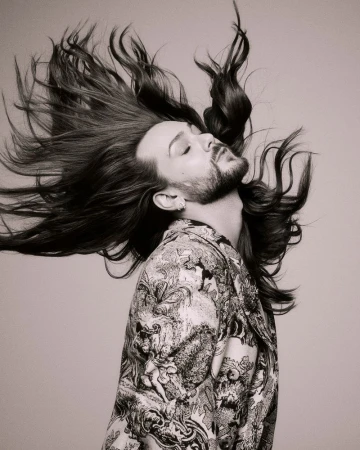 Riccardo Simonetti flipping long dark hair wearing a patterned blazer against a neutral grey studio background