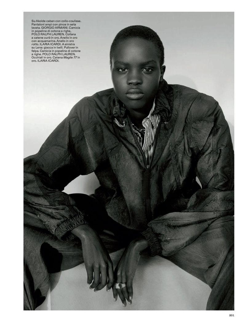 Striking studio portrait of a Black woman in a sporty jacket and casual striped shirt, seated with hands resting, exuding a confident gaze.