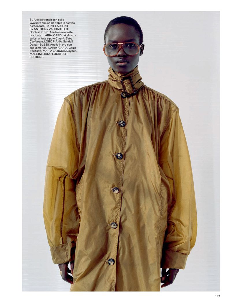 A dark-skinned fashion model poses in a tan trench coat and translucent glasses against a bright white backdrop in a studio setting.
