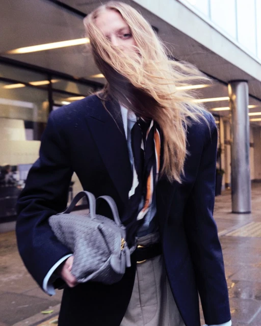 Fashion shot of a blond model in a blazer and light-colored pants, carrying a gray woven purse, hair blowing in the wind, outdoors.